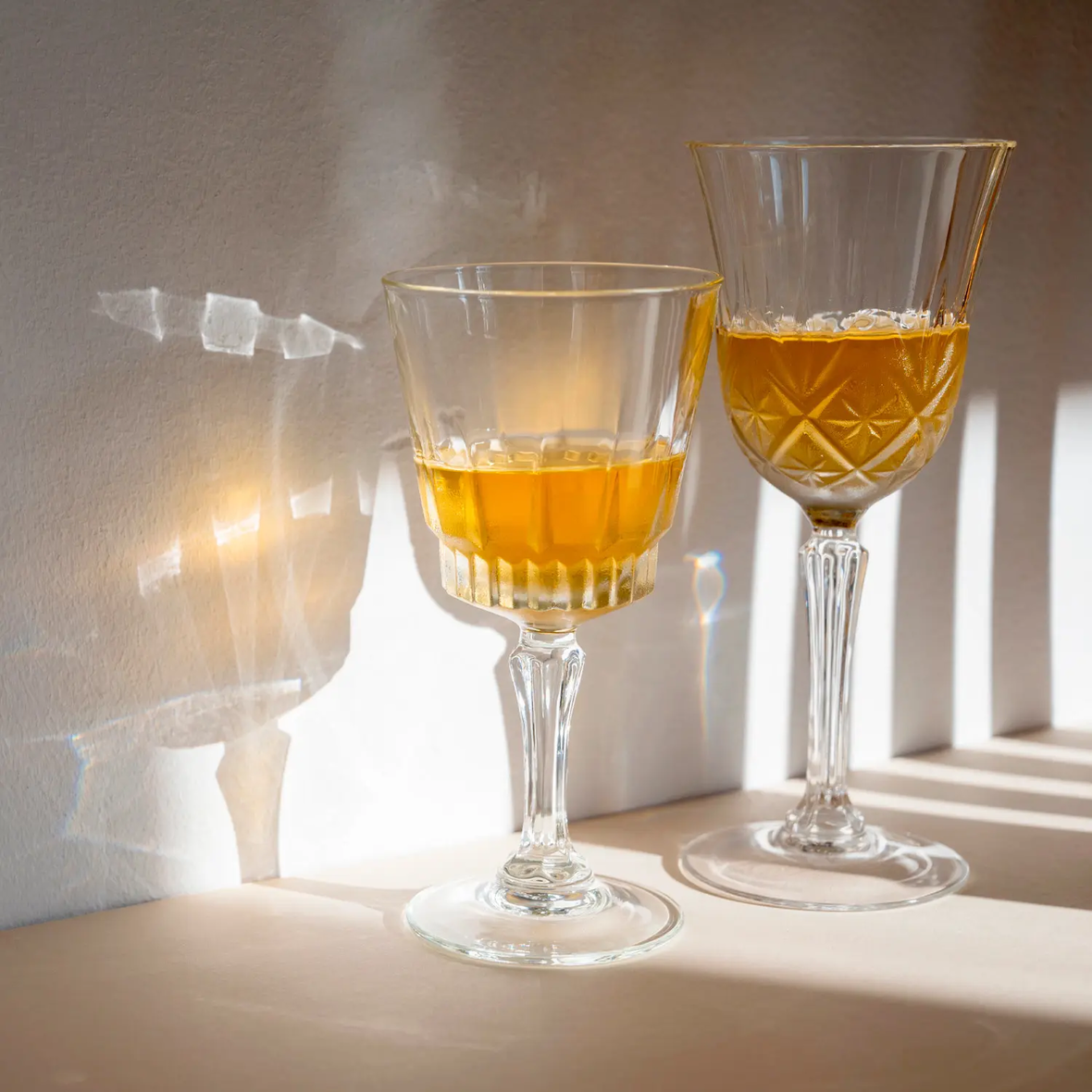 Zwei Kristallgläser, teilweise mit Orangensaft gefüllt, stehen nebeneinander auf einem Tisch im warmen Sonnenlicht. Die Gläser werfen beeindruckende Lichtreflexionen und Schatten an die Wand, während das Licht ihre klaren, detaillierten Oberflächen betont.